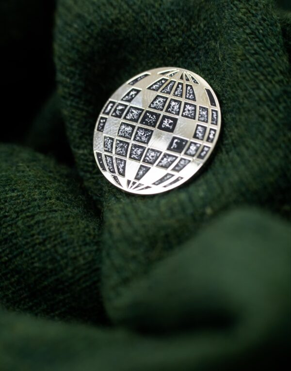 Black round metal pin with silver outlines and filled squares of a disco ball on a dark green sweater