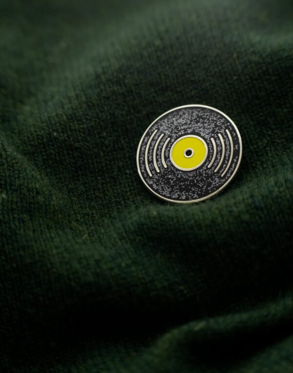 Black round metal pin with silver outline of a vinyl and yellow centre on a dark green sweater
