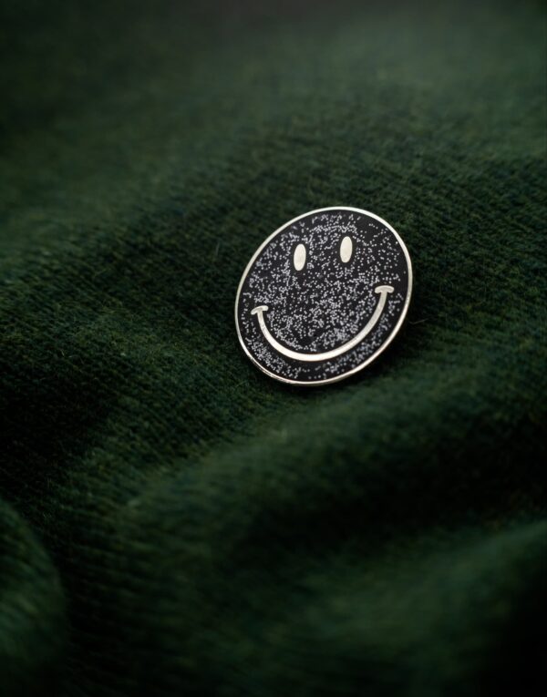 Black round metal pin with silver smiley face and glitter on a dark green sweater